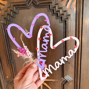 May include: A heart-shaped wooden sign with the word "Mama" in cursive. The sign is in pink and purple, decorated with small heart accents and a small bunch of dried flowers.