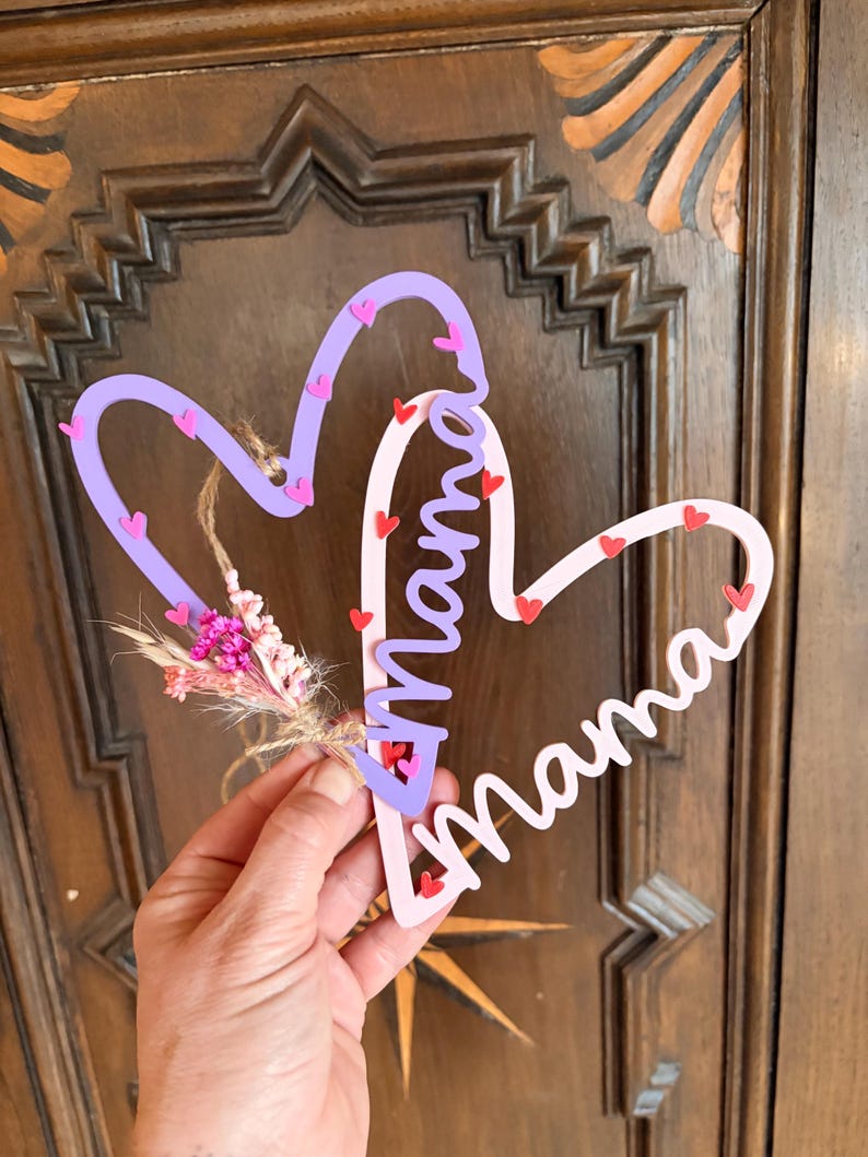 May include: A heart-shaped wooden sign with the word "Mama" in cursive. The sign is in pink and purple, decorated with small heart accents and a small bunch of dried flowers.