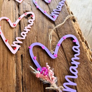 May include: A collection of heart-shaped wooden decorations, each with the word "Mama" written in cursive. The ornaments are in pink and purple hues, decorated with tiny heart details. One has a small dried flower bouquet.