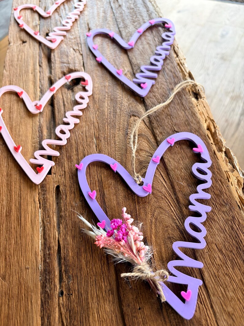 May include: A collection of heart-shaped wooden decorations, each with the word "Mama" written in cursive. The ornaments are in pink and purple hues, decorated with tiny heart details. One has a small dried flower bouquet.