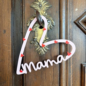 May include: A pink heart-shaped door hanger with the word "mama" in cursive. Small red hearts are scattered along the edges. The hanger is suspended from a decorative brass door handle on a wooden door.
