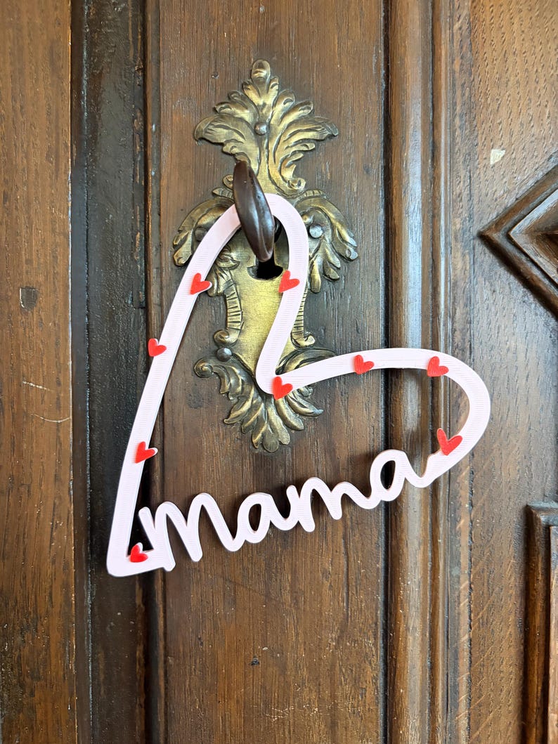 May include: A pink heart-shaped door hanger with the word "mama" in cursive. Small red hearts are scattered along the edges. The hanger is suspended from a decorative brass door handle on a wooden door.