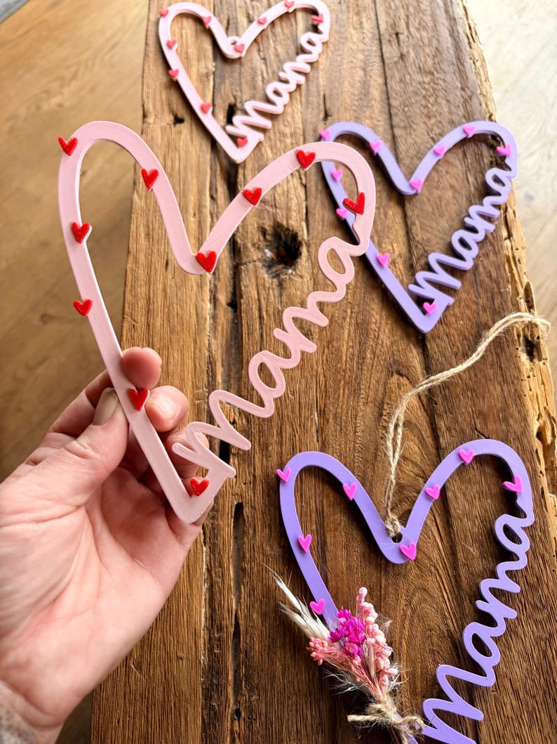 May include: Pink, lilac, and red heart-shaped decorations with the word "Mama" in cursive. Each heart is decorated with small heart accents. One heart has a small bouquet of pink flowers.