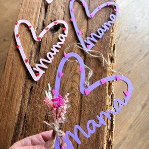 May include: Heart-shaped wooden decorations spelling "Mama" in cursive. The hearts are light pink and purple, decorated with small red and pink hearts. One heart is paired with a small bouquet of dried flowers.