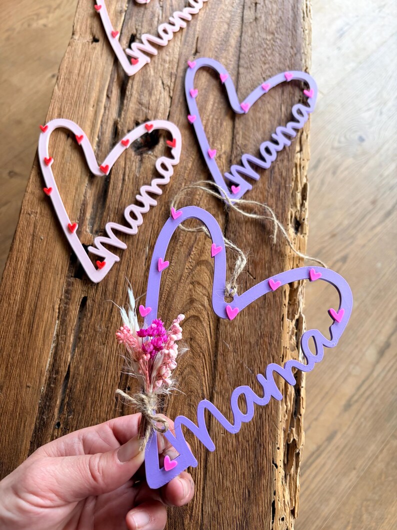 May include: Heart-shaped wooden decorations spelling "Mama" in cursive. The hearts are light pink and purple, decorated with small red and pink hearts. One heart is paired with a small bouquet of dried flowers.