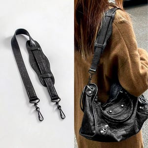 Könnte beinhalten: Ein schwarzer, texturierter Schulterriemen aus Leder mit einem geformten Schulterpolster und zwei drehbaren Karabinerverschlüssen aus Metall an den Enden. Der Riemen ist separat und an einer schwarzen Ledertasche mit silbernen Metallbeschlägen zu sehen.