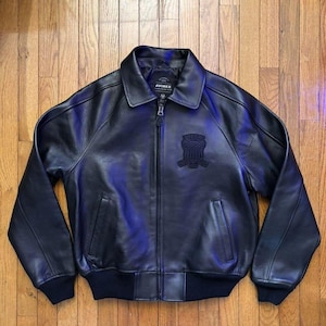 May include: Black leather bomber jacket with a zip-up front, collar, and ribbed cuffs and hem. The jacket features a small emblem on the chest and the brand name "AVIREX" on the label.