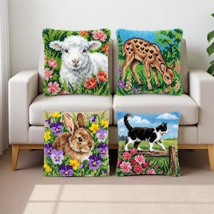 Op de afbeelding: Vier decoratieve kussens met geborduurde dierenontwerpen. De kussens tonen een lam, een hert, een konijn en een zwart-witte kat. Elk kussen is omgeven door kleurrijke bloemen en groen, op een lichtgekleurde bank.