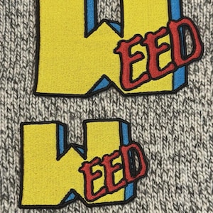 Könnte beinhalten: Zwei gestickte Aufnäher mit dem Wort "WEED" in einer stilisierten Schriftart. Die Aufnäher haben gelbe Buchstaben mit blauen Umrissen und einen roten "EED"-Abschnitt. Die Aufnäher befinden sich auf einem grau-weißen Strickstoffhintergrund.