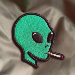 Könnte beinhalten: Gestickter Aufnäher mit einem grünen Alien, das eine Zigarette raucht. Der Alien hat große schwarze Augen und eine schwarze Umrandung. Der Aufnäher befindet sich vor einem neutralen Hintergrund.