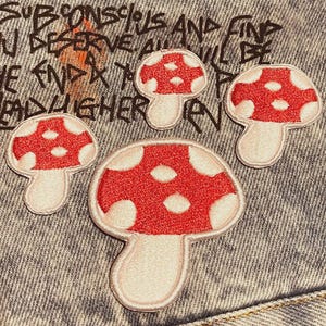 Könnte beinhalten: Vier Pilzaufnäher mit roten Kappen und weißen Punkten sind auf einem Jeanshintergrund angeordnet. Die Aufnäher haben einen weißen Rand und Stiel. Der Hintergrund hat handgeschriebenen Text in Schwarz.