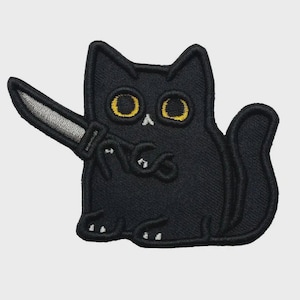 Könnte beinhalten: Gestickter Aufnäher mit einer schwarzen Katze, die ein silbernes Messer hält. Die Katze hat große gelbe Augen und weiße Akzente an Pfoten und Nase. Das Design ist minimalistisch und hat eine spielerische, leicht bedrohliche Ästhetik.