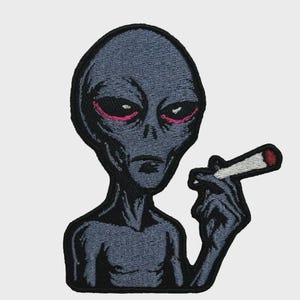 Könnte beinhalten: Gestickter Aufnäher mit einem grauen Alien mit roten Augen, der eine brennende Zigarette hält. Der Alien hat einen großen Kopf und einen schlanken Körper mit schwarzem Rand. Die Zigarette hat einen weißen Filter und eine rote Spitze.