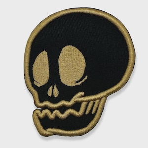 Könnte beinhalten: Gestickter Aufnäher mit einem stilisierten Totenkopf-Design. Der Totenkopf ist schwarz mit goldfarbenen Details, einschließlich Umriss und Gesichtszügen. Der Aufnäher hat einen goldenen Rand, geeignet für Kleidung oder Accessoires.