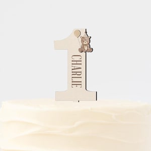May include: A white cake with white frosting on a wooden cake stand. A wooden cake topper with the number one and the name Charlie is on top of the cake.