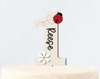 May include: White cake with buttercream frosting topped with a wooden number one cake topper decorated with a ladybug and a daisy. The topper also has the name "Reese" written on it.