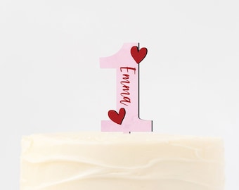Könnte beinhalten: Ein weißer Kuchen mit rosa Zuckerguss und einem rosa und roten Nummer-eins-Kuchentopper mit dem Namen "Emma" darauf.