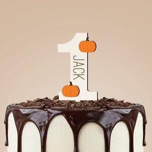 May include: A white cake with chocolate frosting and chocolate shavings on top. A wooden cake topper with the number one and the name "Jack" is on top of the cake. The cake topper also has two small orange pumpkins on it.