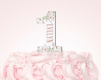 May include: A pink frosted cake with a floral design number one cake topper. The topper has the name "Evelyn" written on it.
