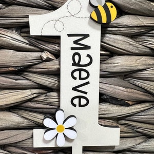 May include: A white wooden number one with a black outline and the name "Maeve" written vertically in black. A yellow and black bee with gray wings is attached to the top of the number. A white daisy with a yellow center is attached to the bottom of the number.
