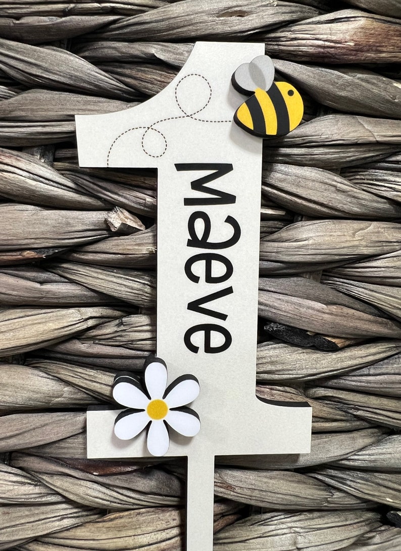 May include: A white wooden number one with a black outline and the name "Maeve" written vertically in black. A yellow and black bee with gray wings is attached to the top of the number. A white daisy with a yellow center is attached to the bottom of the number.
