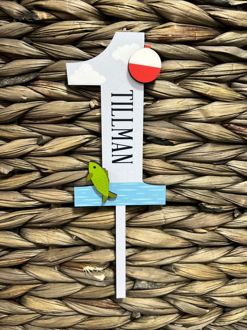 Puede incluir: Un n&uacute;mero uno de madera blanco con un cielo azul y blanco, un flotador de pesca rojo y blanco, un pez verde saltando fuera del agua azul y el nombre "Tillman" escrito verticalmente en el n&uacute;mero.