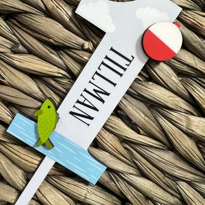Puede incluir: Un n&uacute;mero uno de madera blanco con la palabra "Tillman" escrita verticalmente en &eacute;l. El n&uacute;mero uno tiene un dise&ntilde;o de agua azul y blanco en la parte inferior, un pez verde y una boya roja y blanca.