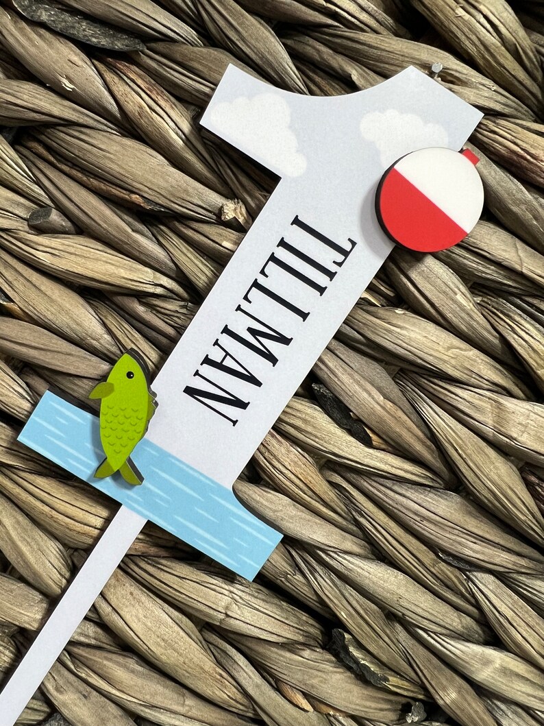 Puede incluir: Un n&uacute;mero uno de madera blanco con la palabra "Tillman" escrita verticalmente en &eacute;l. El n&uacute;mero uno tiene un dise&ntilde;o de agua azul y blanco en la parte inferior, un pez verde y una boya roja y blanca.