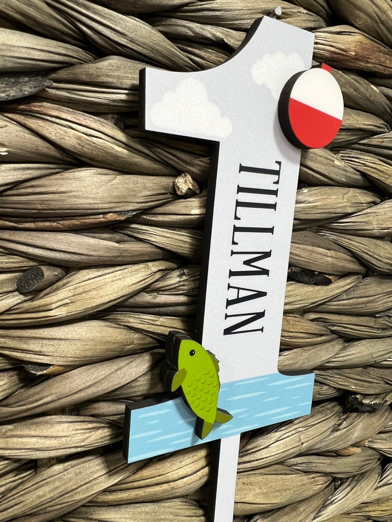 Puede incluir: Un n&uacute;mero uno de madera con un fondo de cielo azul, nubes blancas, un flotador de pesca rojo y blanco, un pez verde saltando fuera del agua azul y el nombre "Tillman" escrito en negro.