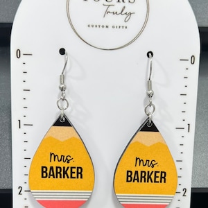 May include: A pair of yellow and red pencil-shaped earrings with the text "mrs. BARKER" on each earring. The earrings are hanging from silver hooks.