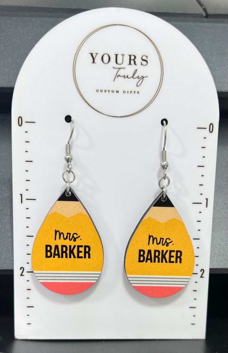 May include: A pair of yellow and red pencil-shaped earrings with the text "mrs. BARKER" on each earring. The earrings are hanging from silver hooks.