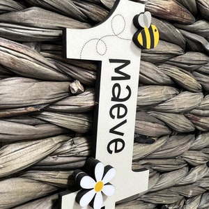 May include: A white wooden number one with a black border. The number is decorated with a black and yellow bee, a white daisy with a yellow center, and the name "Maeve" written in black. The number is leaning against a woven wicker background.