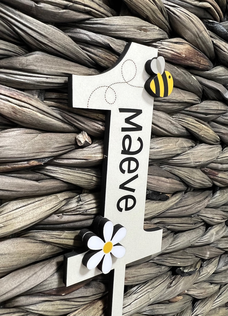 May include: A white wooden number one with a black border. The number is decorated with a black and yellow bee, a white daisy with a yellow center, and the name "Maeve" written in black. The number is leaning against a woven wicker background.