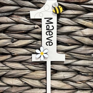 May include: White wooden number one with a black outline and the name "Maeve" written vertically. A yellow and black bee and a white daisy with a yellow center are attached to the number.