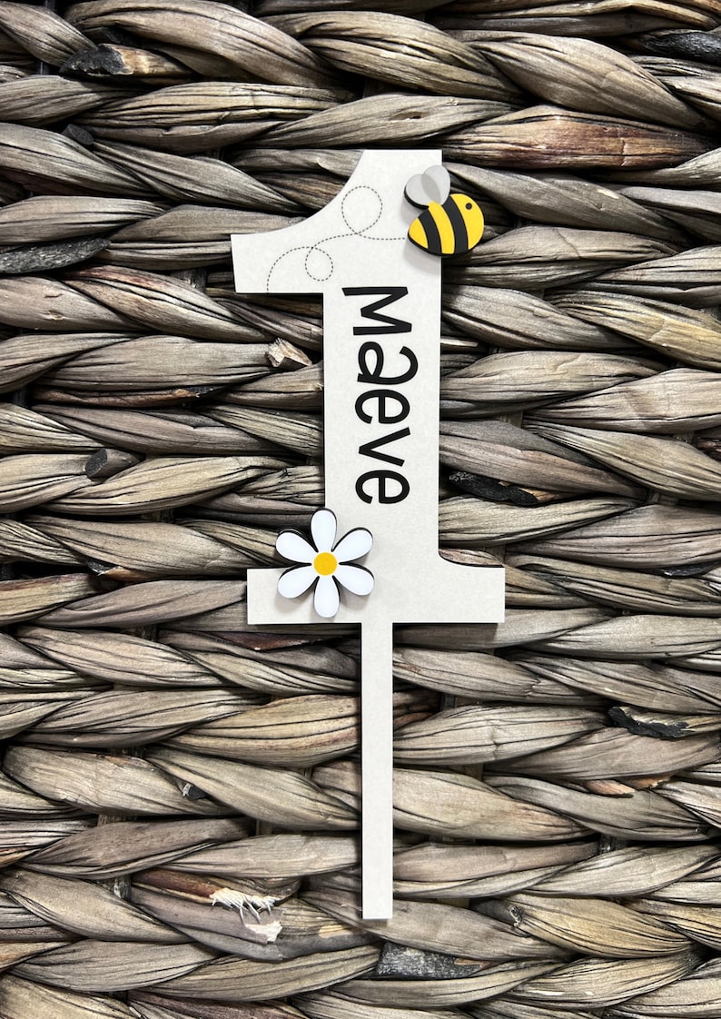 May include: White wooden number one with a black outline and the name "Maeve" written vertically. A yellow and black bee and a white daisy with a yellow center are attached to the number.