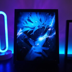 May include: A framed art print with a blue anime-style illustration of a character with spiky hair and a focused expression. The artwork is illuminated with a blue light, creating a dramatic effect. The frame is black.