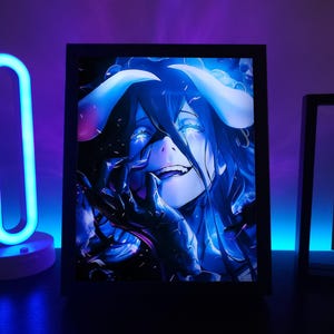 Puede incluir: Una impresión artística enmarcada de anime que presenta un personaje con cabello azul, ojos brillantes y cuernos. La obra de arte es predominantemente azul y negro, con una sonrisa siniestra. El marco es negro y la imagen está iluminada con un brillo azul.