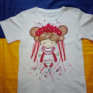 Könnte beinhalten: Weißes T-Shirt mit einer handgemalten Illustration einer Puppe mit rotem Blumenkranz und traditioneller ukrainischer Kleidung. Die Puppe hat braune Haare und geschlossene Augen. Rote und schwarze Farbspritzer umgeben die Puppe.