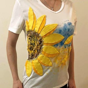 Puede incluir: Camiseta blanca con cuello en V con un gran diseño de girasol. El girasol tiene pétalos amarillos y un centro marrón, adornado con lentejuelas. También hay detalles de acuarela azules y amarillos.