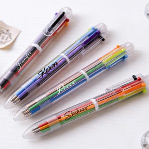 Puede incluir: Cuatro bolígrafos transparentes multicolores con los nombres Jennifer, Karen, Reece y Shahnaz impresos. Cada bolígrafo tiene un cuerpo transparente con varios colores de tinta visibles en el interior. Los bolígrafos están sobre una superficie blanca.