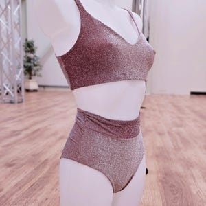 Peut inclure: Ensemble de deux pièces de vêtements de sport rose doré scintillant. Le haut est un soutien-gorge de sport à col rond et le bas est un slip taille haute. Le tissu a une texture scintillante. L'ensemble est présenté sur un mannequin.