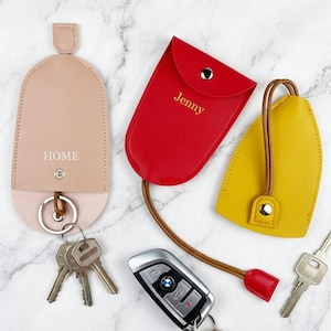 May include: Three leather key holders in beige, red, and yellow. The beige holder has "HOME" printed on it. The red holder is labeled "Jenny". A car key and several metal keys are also present.