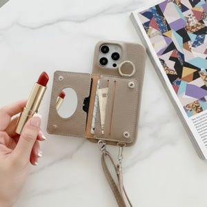 Puede incluir: Una funda de teléfono de cuero color topo con un accesorio de cartera, con un espejo y ranuras para tarjetas. Un lápiz labial rojo en un tubo dorado se sostiene cerca. La funda tiene un soporte de anillo y una correa desmontable.