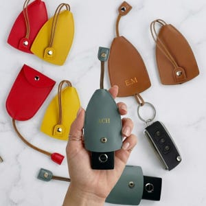 Könnte beinhalten: Leder-Schlüsseletuis in Rot, Gelb, Braun und Teal. Einige sind mit Initialen personalisiert. Jedes hat einen Riemen und einen Druckknopfverschluss. Die Schlüsseletuis schützen die Schlüssel.