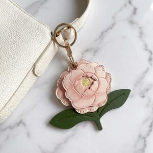 Könnte beinhalten: Ein rosafarbener Lederblumen-Schlüsselanhänger mit einem goldfarbenen Verschluss und Ring. Die Blume hat eine gelbe Mitte und ist an einem grünen Blatt befestigt. Der Schlüsselanhänger befindet sich neben einer weißen Handtasche.