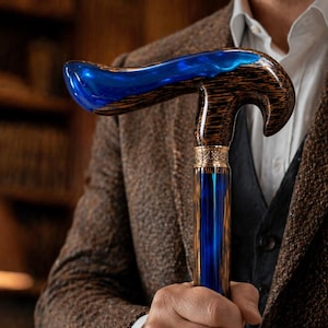 Könnte beinhalten: Handgefertigter Gehstock mit gebogenem Griff und blauem Harzaufsatz. Der Schaft ist aus dunklem Holz mit blauem Kern, akzentuiert durch goldfarbene Metallbänder. Der Stock wird von einer Person in einer braunen Tweedjacke gehalten.