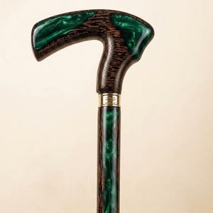 Puede incluir: Bastón de madera con un mango curvo y un eje. El mango y el eje presentan un patrón de vetas de madera oscura con una incrustación de resina verde vibrante. Una banda dorada acentúa la unión entre el mango y el eje.