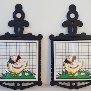 Puede incluir: Dos posavasos de hierro fundido negro con azulejos de cerámica blanca que representan una gallina y pollitos de dibujos animados. Los azulejos tienen un patrón de cuadros.
