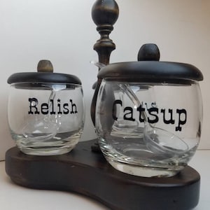 May include: A wooden condiment holder with two glass jars, one labeled "Relish" and the other "Catsup". Each jar has a small spoon inside.
