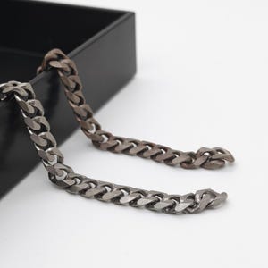 May include: Two metal chain necklaces with a curb link design. One chain is silver, and the other is a dark bronze color. The chains are draped over the edge of a black box against a white background.